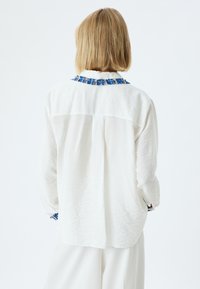 Camisa blanca de manga larga con un tejido texturizado y detalle de cuello azul deshilachado. Presenta una sola costura en la parte espalda y puños con acentos estampados.
