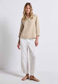 Beige, strukturierter Langarmoberteil mit V-Ausschnitt und Knopfverschluss vorn. Kombiniert mit locker sitzenden weißen Hosen und braunen Sandalen.