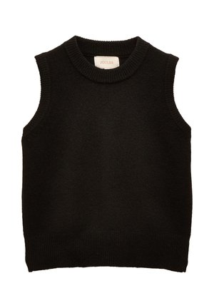 Junge Frau trägt schwarzen ärmellosen Pullover über weißer Bluse, blaue hochgekrempelte Jeans und braune Loafer, steht vor beigem Hintergrund.