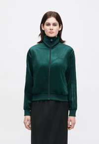 Green velvet zip-up jacket with a high collar, featuring embroidered logo and stripes along sleeves, paired with a black skirt.