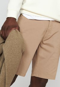 Beige bavlněné šortky s plochým předním dílem a postranními kapsami. Osoba drží béžový svetr a má na sobě světle zbarvenou mikinu.