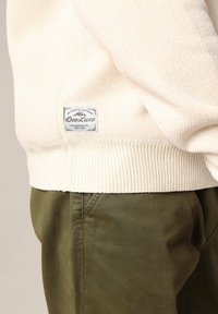 Crèmekleurige gebreide trui met geribbeld uiteinde, voorzien van een geweven label met de branding "DeeLuxe". Gecombineerd met olijfgroene textuurbroek.