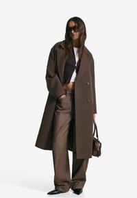 Femme portant un manteau brun oversize, un pantalon large brun, un crop top, de grandes lunettes de soleil, des chaussures noires à bouts pointus et tenant un sac à main marron.