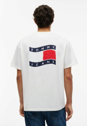 Mann von hinten gesehen, trägt ein weißes T-Shirt mit rot-weiß-blauem Tommy Jeans-Logo und blaue Jeans.