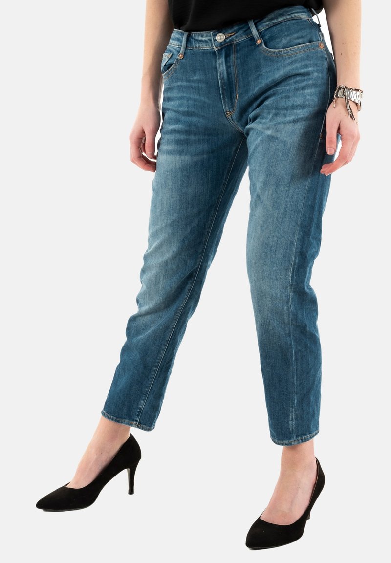 Jean en denim bleu avec une coupe ajustée, doté de cinq poches, d'un bouton et d'une fermeture éclair, avec un délavage légèrement usé. Porté avec des talons hauts noirs.