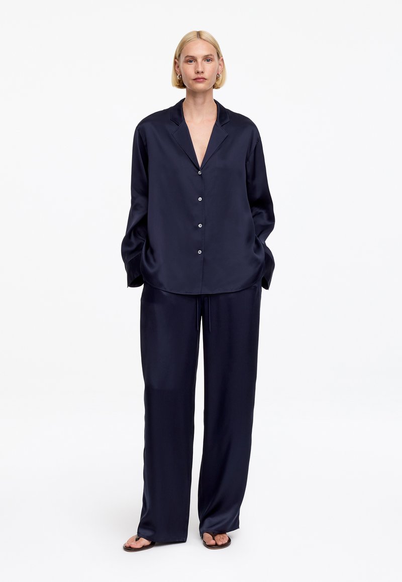 Femme aux cheveux blonds courts portant une chemise ample en satin bleu marine boutonnée et un pantalon large, debout les mains dans les poches devant un fond blanc.