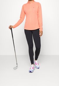 Haut corail vif à manches longues avec fermeture éclair, en tissu texturé, associé à des leggings noirs et des chaussures de sport multicolores. Club de golf tenu en main.
