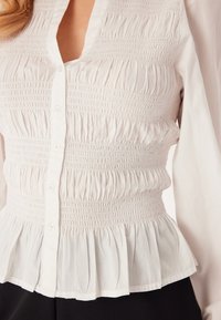 Hvid bluse med smock-struktur og flared kant, der har en delvis knaplukning og blødt, let stof. Lange ærmer smalner let ind.