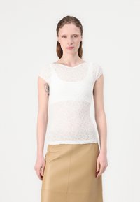 Top alb din dantelă cu mâneci scurte, având un model floral și o textură semi-transparentă. Purtați peste o fustă din piele bej.