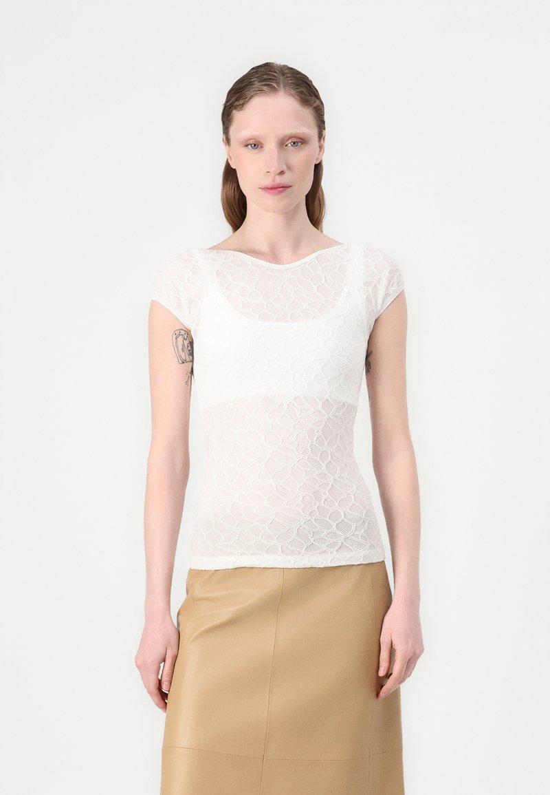 Top alb din dantelă cu mâneci scurte, având un model floral și o textură semi-transparentă. Purtați peste o fustă din piele bej.
