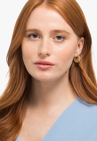 Boucles d'oreilles en or avec des designs découpés complexes, présentant une texture lisse. L'arrière-plan est légèrement flou, mettant en valeur les boucles d'oreilles.