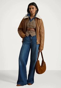 Žena v hnedej koženej bunde s ovčím golierom, opásanom béžovým vestom, modrých riflových širokých nohaviciach, s hnedou zamšovou kabelkou.