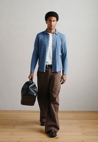 Camisa azul com botões por cima de uma t-shirt branca, calças largas castanhas, mala preta com padrão texturizado e sapatos pretos. Fundo de pavimento em madeira lisa.