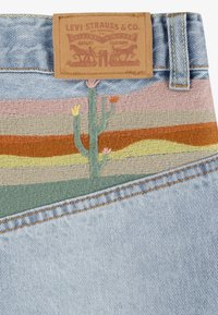 Denim szövet barackszínből készült bőrpótlékkal; sokszínű hímzés díszíti, amely egy kaktuszt és sivatagi tájat ábrázol rózsaszín, narancs és zöld árnyalataiban.