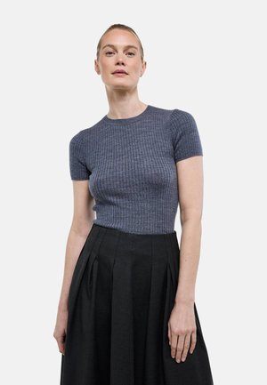 Femme aux cheveux blonds attachés portant un haut gris côtelé à manches courtes et une jupe plissée noire taille haute, de face.