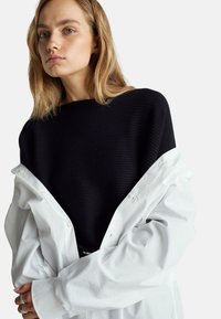 Czarny sweter z prążkowanym wzorem w połączeniu z luźną białą koszulą zapinaną na guziki, ukazując kontrastujące kolory i faktury. Model pozuje z założonymi na siebie rękami.