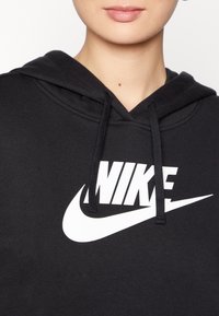 Person klädd i en svart Nike-hoodie med vitt Nike-logo och dragsnören, som visar hals och läppar mot en enfärgad bakgrund.