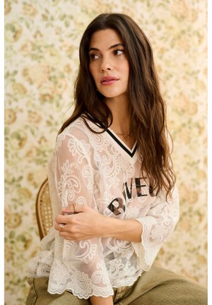 Hosszú sötét hajú nő ül egy széken, fehér csipke felsőt visel fekete szöveggel egy virágmintás háttéren.