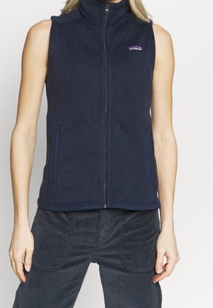 Gilet sans manches en polaire bleu marine zippé porté par une personne à la peau claire et aux cheveux blonds, associé à un pantalon en velours côtelé bleu marine foncé.