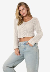 Ljusbeige kortstickad tröja med V-ringning och öppen lattédesign. Har långa ärmar med utsvängda manschetter. Bärs med högmidjade blåjeans.