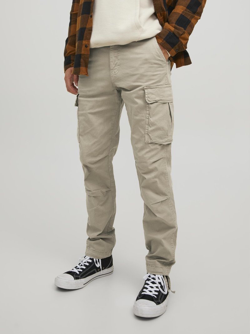 Beige cargo pants with side pockets, made of cotton fabric. Model wears black and white sneakers, paired with a plaid shirt.