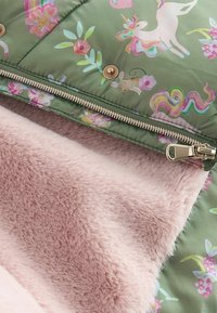 Tissu matelassé vert avec imprimés de licornes et de lapins, boutons-pression dorés, fermeture éclair partiellement ouverte pour montrer une doublure en fausse fourrure rose douce.
