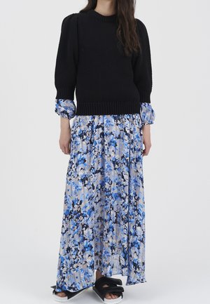 Kobieta ubrana w czarny dzianinowy sweter na niebieskiej, białej i czarnej kwiecistej sukience maxi, do tego czarne sandały na grube paski z odkrytymi palcami.