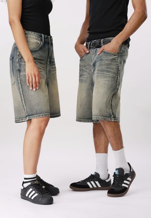To personer iført sorte toppe, falmede denimshorts, hvide sokker og sorte Adidas-sneakers, der står overfor hinanden på en ensfarvet baggrund.