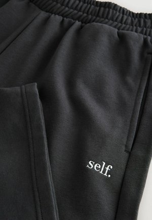 Pantaloni jogger neri con vita elasticizzata, caratterizzati da una texture morbida e un logo ricamato bianco "self." vicino alla tasca.