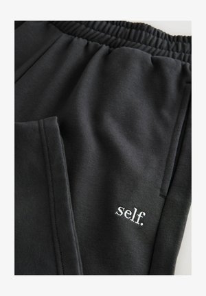 Crne hlače za trčanje s elastičnim pojasom, s mekanom teksturom i bijelim izvezanim logotipom "self." blizu džepa.