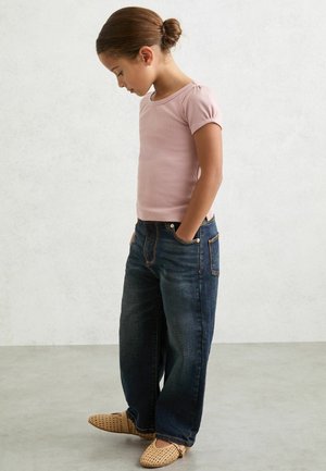 Světlá růžová žebrovaná košile s krátkým rukávem, tmavě modré široké džíny, béžové pletené nazouvací boty. Dítě stojí s rukama v kapsách, hlava skloněná dolů.