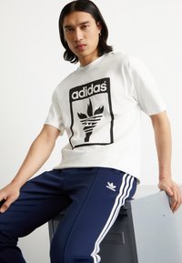 White cotton t-shirt featuring a black graphic logo with a stylized torch design. Paired with navy athletic pants with white stripes.