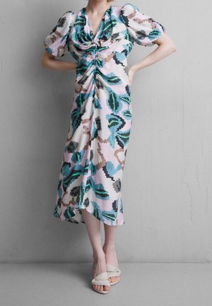 Kobieta stoi z rękami na biodrach, ubrana w sukienkę midi z bufiastymi rękawami i różowo-zielono-niebieskim abstrakcyjnym kwiatowym wzorem.