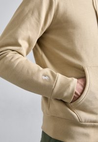 Beiger Sweatshirtärmel mit weiß gesticktem Logo nahe dem Handgelenk und Hand, die in die passende Vordertasche gesteckt ist.