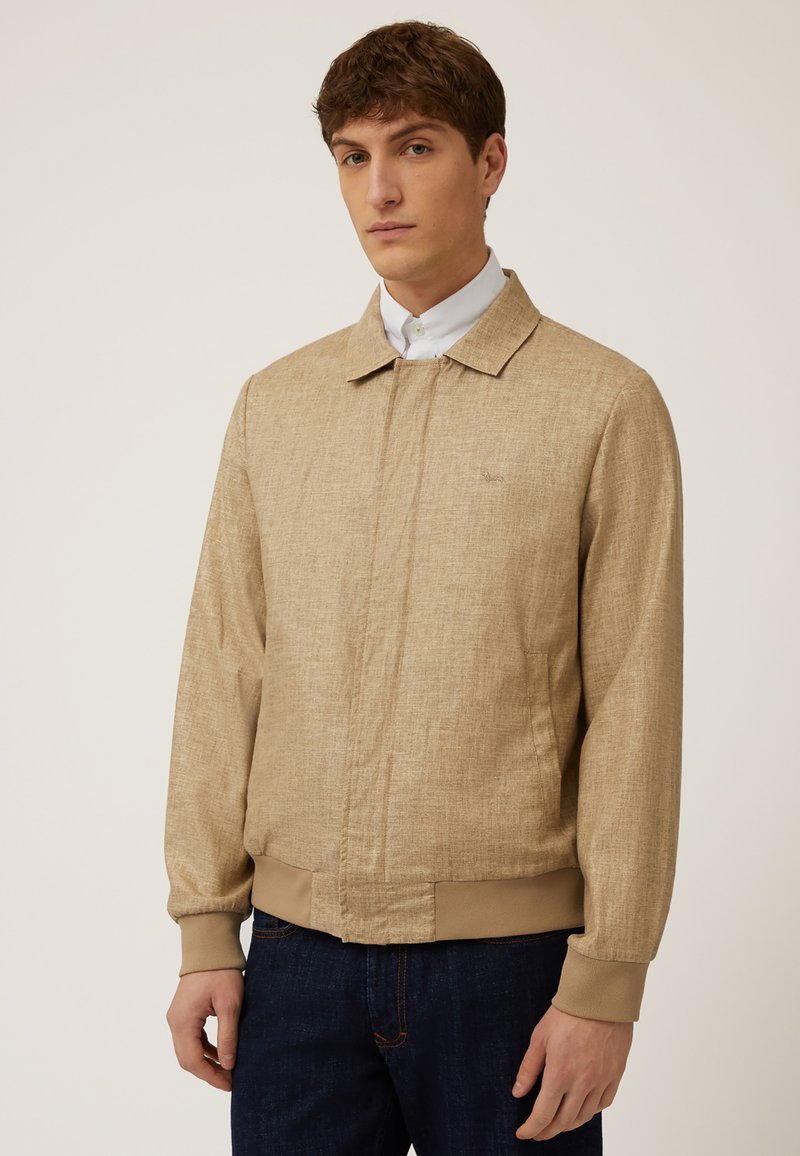 Chaqueta bomber ligera en tono beige con un acabado texturizado, puños y dobladillo acanalados, y cierre de cremallera. Cuenta con un cuello clásico y dos bolsillos laterales.