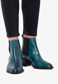 Botines de cuero color verde azulado con paneles laterales elásticos, punta puntiaguda y tacón bajo de bloque, combinados con jeans ajustados negros.