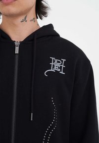 Sudadera con cremallera negra, con un logo plateado en el pecho, adornos de puntos a lo largo de un lado y cordones para ajuste.