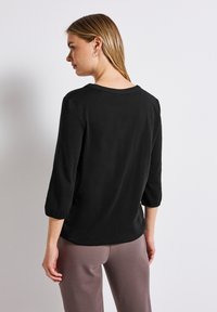 Zwarte top met driekwartmouwen en een ontspannen pasvorm. Heeft een ronde halslijn en geplooide details bij de mouwmanchetten. Soepele stoftextuur.