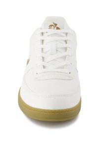 Sneaker bianco con tomaia in pelle liscia, semplice chiusura con lacci e suola in gomma marrone con dettagli testurizzati. Logo dorato sul lato.