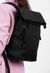 Černý látkový batoh s rolovacím uzávěrem, nastavitelnými popruhy a dvěma bočními zipy. Hladká textura a minimalistické designové prvky.