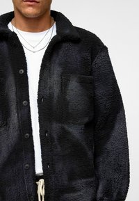 Zwarte fleece shirtjack met een knoopsluiting aan de voorkant, gestructureerd oppervlak, puntkraag en een borstzak. Draagt over een wit T-shirt.