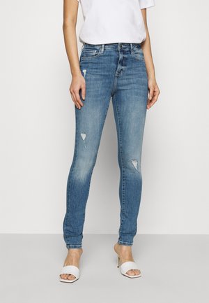 Woman wearing light blue ripped skinny jeans, white short-sleeve shirt, and white open-toe heeled sandals standing against a plain background.