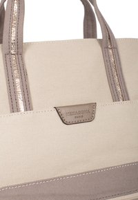 Beige stoffen tote bag met taupe accenten, dubbele handvatten en een textuurlogo-patch. Reflecterende stiksel voegt detail aan het ontwerp toe.