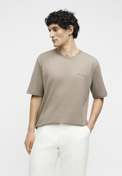 Άνδρας με σγουρά σκουρόχρωμα μαλλιά που φοράει μπεζ κοντομάνικο μπλουζάκι και λευκό παντελόνι, κοιτάζει αριστερά με το ένα χέρι στην τσέπη, σε απλό φόντο.