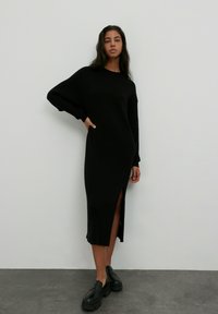 Schwarzes geripptes Strickmidikleid mit langen Ärmeln und seitlichem Schlitz. Kombiniert mit klobigen schwarzen Schuhen. Glatte Textur, minimalistisches Design.