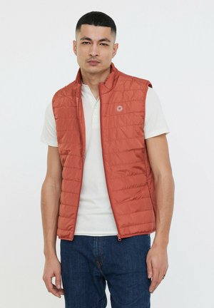 Gilet matelassé rouge avec un col montant, fermeture éclair et emblème logo sur la poitrine gauche. Porté par-dessus une chemise polo blanche et un jean en denim.