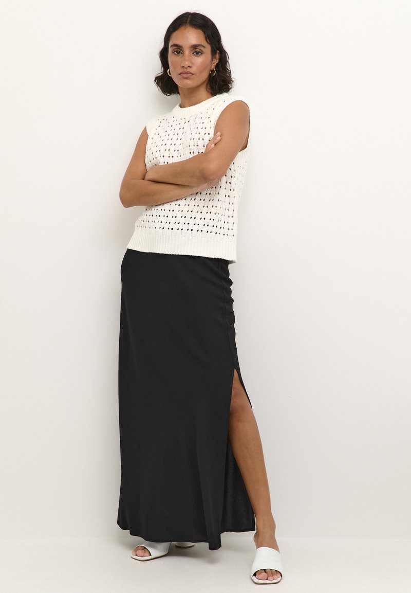 Weiße gestrickte ärmellose Bluse mit offenem Muster, kombiniert mit einem langen schwarzen Rock mit einem seitlichen Schlitz. Das Model trägt weiße Sandalen mit offenen Zehen.