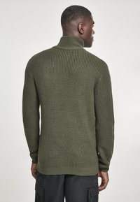 Suéter verde de punto con cuello alto, patrón texturizado y corte holgado, con mangas largas y un dobladillo que se extiende más allá de la cintura.