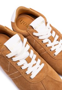 Brown suede sneakers with white laces. Features a stitched design and gold accents on the tongue. Textured insoles visible inside.