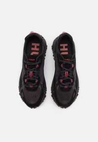Chaussures de sport noires avec une tige en mesh et matériaux synthétiques, accents rouges et semelles texturées. Équipées de lacets noirs et du logo "HUGO" bien visible.